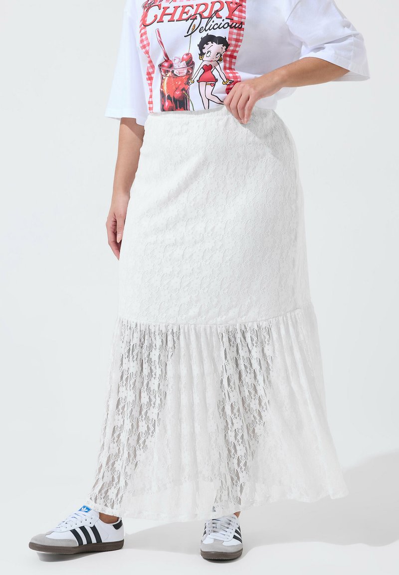 Mujer con una falda larga blanca de encaje, camiseta gráfica roja y blanca, y zapatillas blancas con rayas negras, de pie con una mano en la cintura.