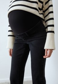 Gravid person klädd i svarta gravidsbyxor och en krämfärgad tröja med svarta ränder, stående mot en vit bakgrund.