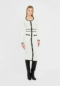 Lang, tætsiddende hvid kjole med sorte detaljer. Har rund halsudskæring, knaplukning foran og to forlommer med guldknapper. Tekstureret stof.