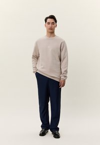 Mladý muž stojaci s rukami v vreckách, oblečený v béžovej mikine, námorníckych modrých nohaviciach a čiernych topánkach na jednoliatom svetlom pozadí.