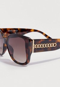 Γυαλιά ηλίου με καφετί φακούς διαβάθμισης, μεγάλα τετράγωνα πλαίσια σε σχέδιο χελώνας και χρυσή αλυσίδα στο πλαϊνό μέρος για επιπλέον λεπτομέρεια.