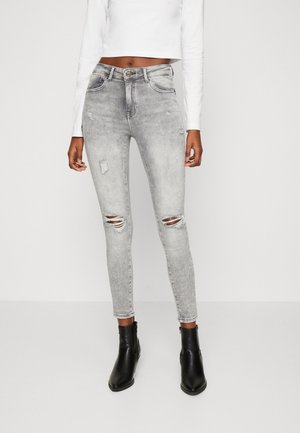 Femme portant un jean skinny taille haute déchiré gris clair, des bottines noires et un top court blanc à manches longues devant un fond uni.