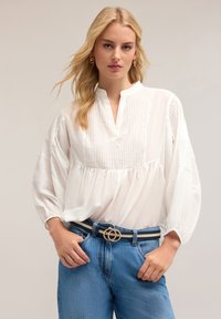 Bluză albă cu butoniere și mâneci bufante, cu dungi verticale. Asortată cu blugi albaștri și o curea neagră cu detalii din aur.