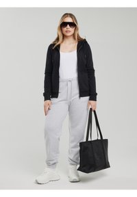 Femeie purtând ochelari de soare, hanorac negru cu fermoar, maiou alb, pantaloni de trening gri, adidași albi și ținând o geantă tote neagră.