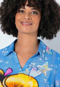 Femme souriante avec des cheveux bouclés portant une chemise bleue ornée d'étoiles de mer colorées, de cœurs et de motifs abstraits sur un fond clair.