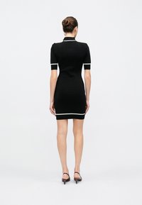 Γυναίκα που στέκεται με την πλάτη στην κάμερα, φοράει εφαρμοστό μαύρο φόρεμα με λευκή επένδυση και καφέ χαμηλά παπούτσια slingback, τα μαλλιά της είναι κότσος.