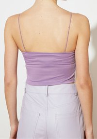 Top crop in lavanda con spalline sottili, texture liscia, forma aderente, abbinato a pantaloni a vita alta di un lieve viola con tasche e cuciture visibili.