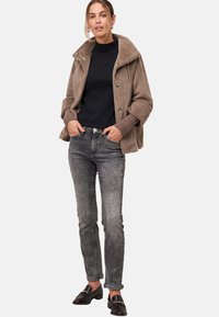 Ruskea, karvainen päällysvaate, jossa on leveä kaulus, musta poolopaita ja harmaat farkut. Mallilla tummat loaferit, kädet taskuissa, rento istuvuus.