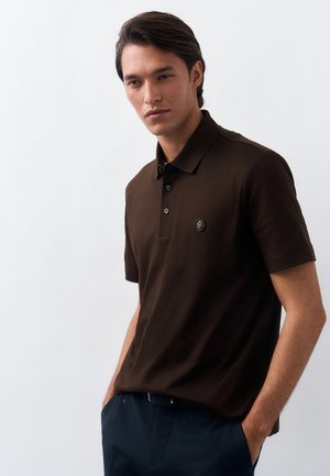 Άνδρας με σκούρα μαλλιά, φοράει καφέ κοντομάνικη πόλο μπλούζα και σκούρο παντελόνι, στέκεται με τα χέρια στις τσέπες μπροστά σε ανοιχτό φόντο.