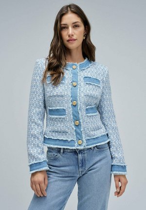 Veste en tweed bleu clair avec des bords effilochés, boutons dorés et accents en denim ; elle possède des poches à patte et un design ajusté.