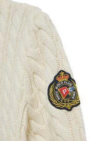 Krämmfärgad stickad tröja med kabelstickningsmönster och en broderad logotypapplikation på axeln. Mjuk textur, avslappnad design.