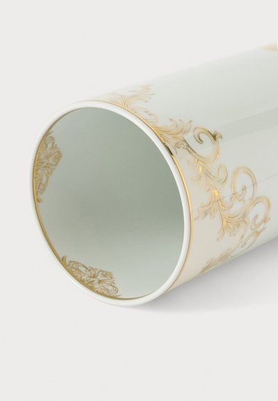 Vase en céramique blanche posé sur le côté avec des arabesques florales dorées détaillées le long du bord et du corps sur un fond blanc.