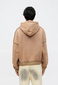 Ruda gobtuvė su gobtuvu, laisvo silueto, minkštu audiniu, nuleistais pečiais ir susmulkintomis kraštinėmis, derinama su šviesios spalvos kelnėmis.