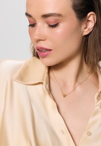 Złoty naszyjnik z wiszącym sercem na delikatnym łańcuszku, noszony z jasnobeżową satynową koszulą z wywiniętym kołnierzykiem i guzikami.