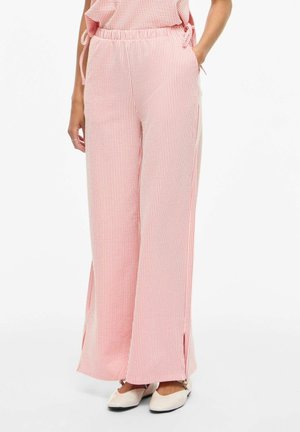 Femeie purtând pantaloni largi cu talie înaltă, cu dungi roz și albe, cu buzunare laterale și pantofi albi fără toc, stând pe un fundal alb.