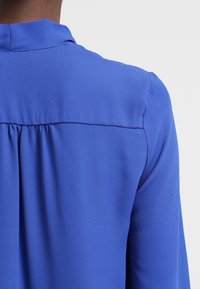 Blå blus med en mjuk, lättvikts tyg. Baksidan har en samlad detalj under halsringningen och lätt utsvängda ärmar.