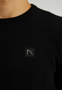 Svart långärmad skjorta i bomull med rund halsringning. Har en liten fyrkantig logotyp med vit söm på bröstet. Slät textur.