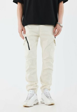 Pantalones cargo beige con dos bolsillos laterales, diseño ajustado, puños elásticos y detalles en cremallera negra. Llevados con zapatillas blancas.