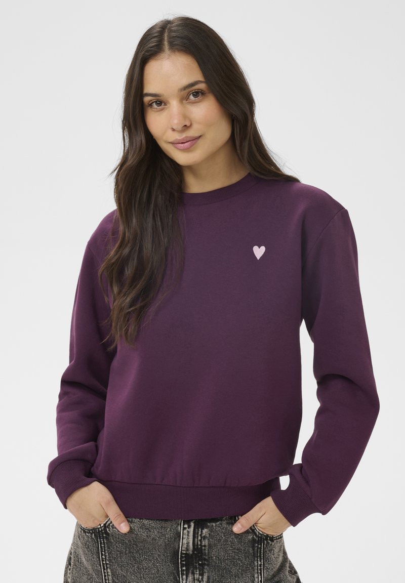 Mladá žena s dlhými tmavými vlasmi má na sebe fialovú mikinu s malým ružovým srdcom na prsiach a čierne rifle s efektom acid wash.