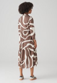 Brown and white patterned dress with long sleeves and a tiered hem. Features a rounded collar and relaxed fit. Worn with casual sneakers.