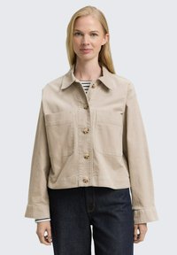 Beige cropped jacket in cotton canvas with a collar, front buttons, and two large pockets. Worn over a black-and-white striped shirt.