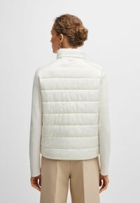 Gilet bianco trapuntato con colletto alto, realizzato in materiale liscio, con cuciture orizzontali e un logo discreto sulla schiena. Abbinato a pantaloni neutri.