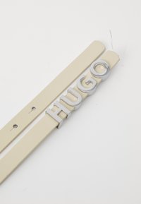 Cintura in pelle beige con una fibbia metallica argento con la scritta "HUGO" in lettere grasse e arrotondate. Due fori di regolazione visibili.