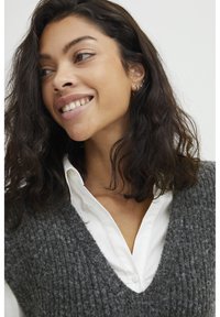 Gilet grigio lavorato a maglia con un motivo testurizzato, caratterizzato da un design a scollo a V. Sotto, è visibile una camicia bianca con colletto.