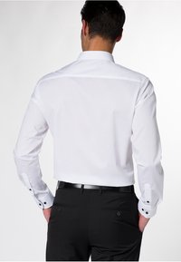 Camicia bianca con maniche lunghe e dettagli in nero sui polsini, indossata con pantaloni neri e cinturino in pelle, che mostra una texture liscia.