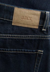 Tmavě modré džíny z denimu s hnědou koženou nášivkou s vyraženým označením "JJXX" nad zadní kapsou a kontrastním šitím.