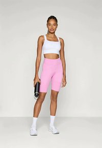 Žena v bielom crop top a ružových cyklistických šortkách, s bielymi ponožkami a teniskami, drží čiernu fľašu na vodu a stojí na jednolatom pozadí.