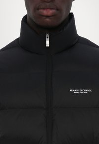 Black puffer jacket with high collar, front zipper, and logo detail. Features a quilted pattern, smooth texture, and minimalistic design.