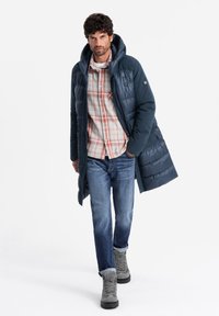 Navyblauer Puffer-Mantel mit Kapuze, ausgestattet mit einem gesteppten Design und Druckknöpfen. Getragen über einem karierten Hemd, kombiniert mit blauen Jeans und grauen Stiefeln.
