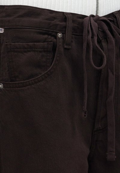 Κοντινό πλάνο σκούρου καφέ τζιν παντελονιού με μπροστινή τσέπη, θηλιά για ζώνη, μεταλλικά καρφιά και κορδόνι στο περιλαίμιο.