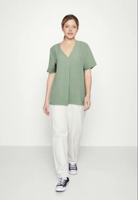 Top verde-claro de manga curta com design em V, apresentando um tecido texturizado. Combinado com calças largas brancas e ténis pretos.