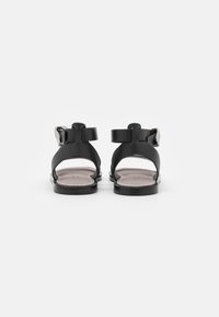 Sandali neri in pelle con retro aperto, cinturino regolabile alla caviglia, design minimalista e suola piatta, caratterizzati da una texture liscia e hardware semplice.