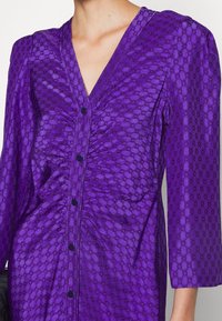 Lila klänning med ett texturerat mönster av sammankopplade cirklar, V-hals, knäppning fram och trekvartslånga ärmar. Rynkning vid midjan.