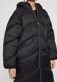 Svart puffjacka med en chevronmönster, långa ärmar, huva och dragkedja hela vägen ner. Tillverkad av ett glänsande, vadderat material.