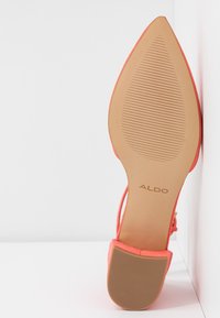 Semelle d'une chaussure plate corail à bout pointu avec texture rainurée et nom de la marque "ALDO" visible, inclinée contre un fond blanc d'angle.
