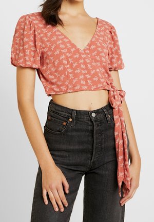 Kvinde iført en pink blomsterwrap crop top med pufærmer og højtaljede sorte jeans mod en lys baggrund.