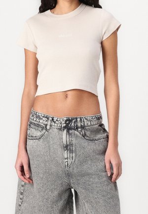 Frau trägt beige, kurzärmeliges, gekürztes Oberteil und hoch taillierte, acid-washed graue Jeans, steht mit entspannten Armen seitlich am Körper.