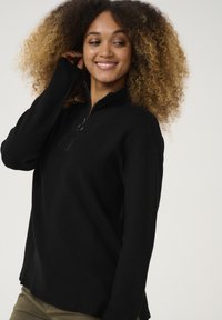Schwarzer Pullover in Rippstepp-Optik mit einem Viertel-Reißverschluss am Hals, seitlichen Schlitzen am Saum und langen Ärmeln.
