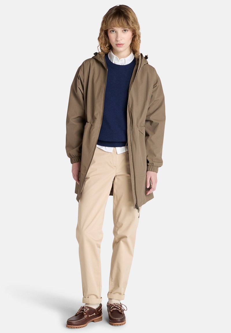 Brązowa kurtka z kapturem i elastycznymi mankietami, noszona na granatowym swetrze, w połączeniu z khaki spodniami i brązowymi butami typu boat shoes, stojąca na białym tle.