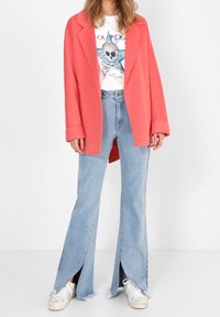 Abrigo grande de mezcla de lana color coral, camiseta gráfica blanca con diseño de calavera, jeans acampanados azul claro con aberturas laterales y zapatillas blancas.
