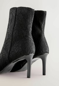 Černé kotníkové boty vyrobené z krajkové tkaniny, s vysokým stiletto podpatkem. Design zahrnuje květinové vzory a hladkou černou podrážku.