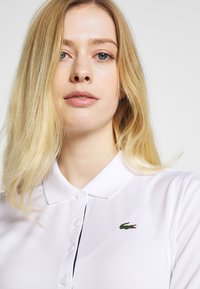 Biała koszulka polo z klasycznym kołnierzykiem, z małym zielonym logo Lacoste na klatce piersiowej. Gładki materiał i szczegóły z zapięciem na guziki.