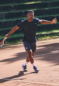 Αθλητικός άνδρας με μπλέ μπλούζα και σορτσάκι, κρατώντας ένα ρακέτα του τένις, σε χωμάτινη γήπεδο με γρασίδι. Τα παπούτσια είναι γκρι με πορτοκαλί λεπτομέρειες.