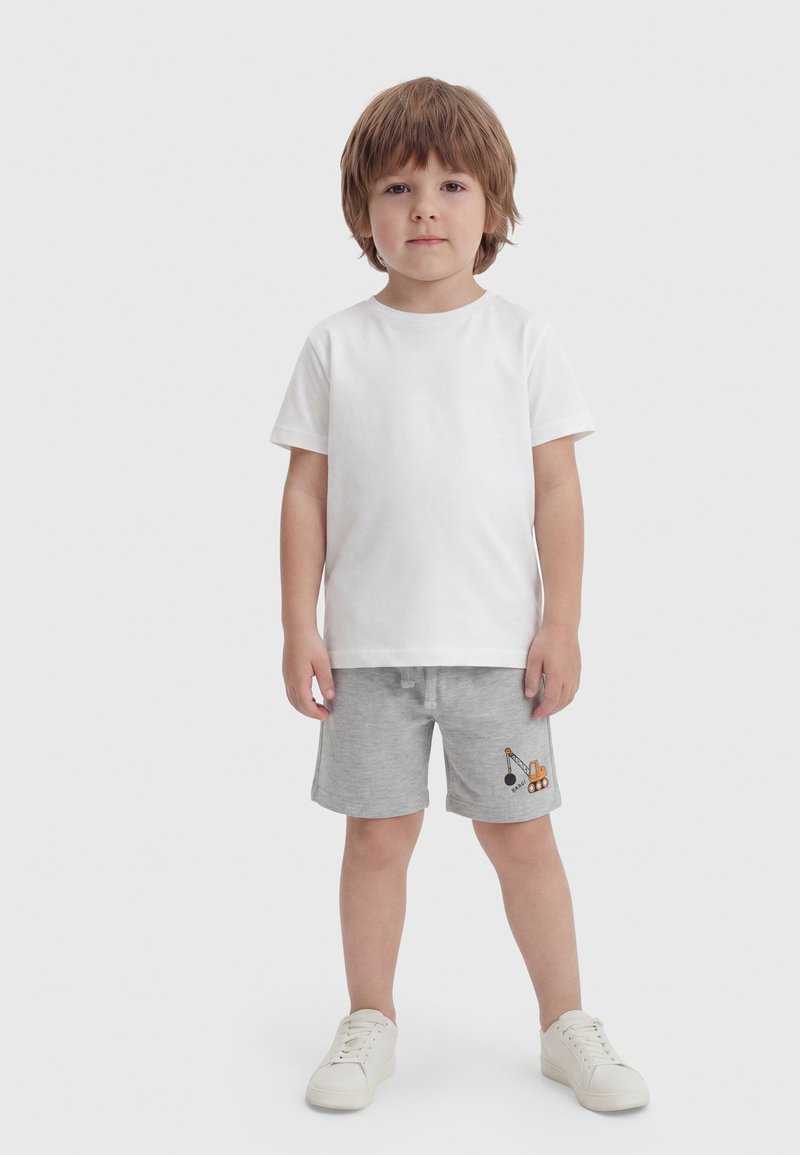 Jeune garçon debout, face à nous, portant un t-shirt blanc uni, un short gris avec un motif de véhicule de construction et des baskets blanches.