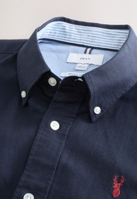 Marineblauw overhemd met knopen en witte knopen, met een rood geborduurd hert op de borst en een lichtblauw gestreept patroon aan de binnenkant van de kraag.