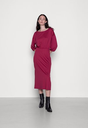 Vestido midi bordeaux com um corpo solto, cintura franzida e mangas longas. Combinado com botas pretas até ao tornozelo. Textura de tecido suave.